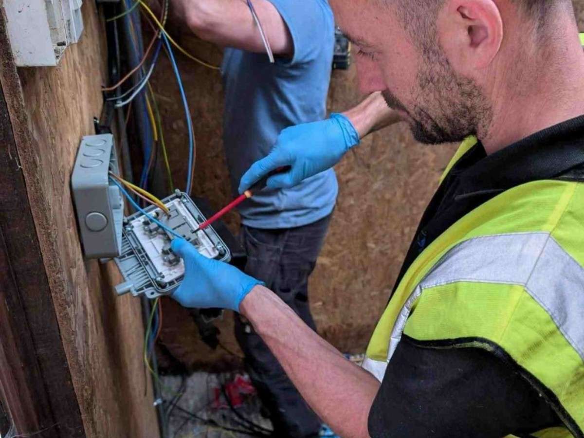 An Electrust Electrical technician in a high-visibility vest and blue gloves, using a screwdriver to work on a junction box mounted on a wooden wall.