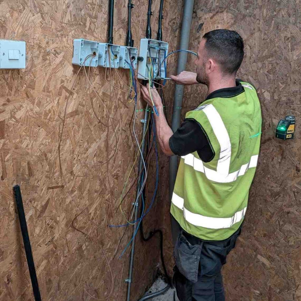 An electrician in a high-visibility vest working on a set of electrical boxes mounted on a wooden wall, with various wires protruding from the boxes.