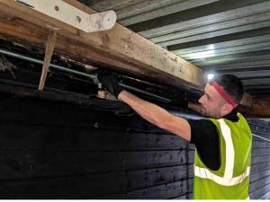 An Electrust Electrical technician wearing a high-visibility vest and a headlamp, adjusting or inspecting a pipe or conduit attached to a wooden beam within a metal structure.