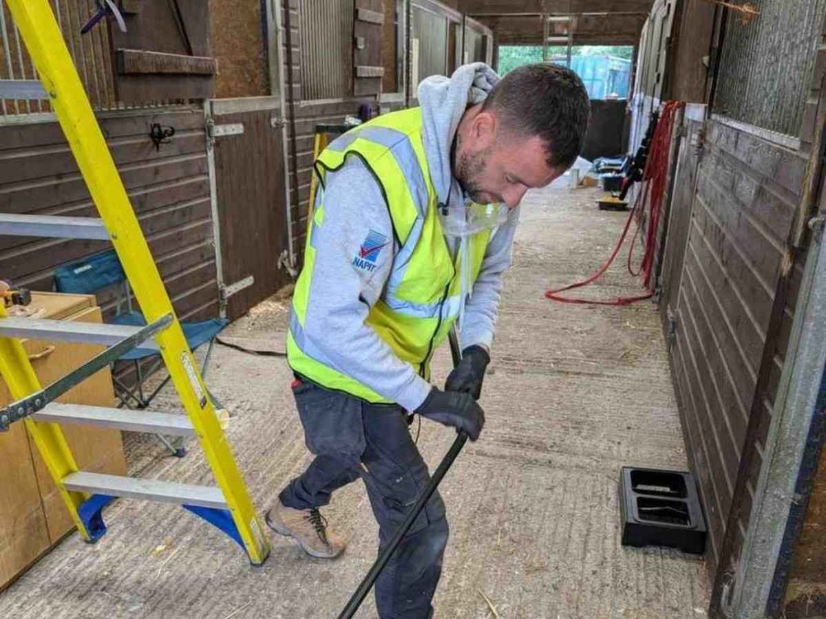 An Electrust Electrical technician in a high-visibility vest is working inside a stable or barn, holding a long black rod or pipe near a yellow ladder.